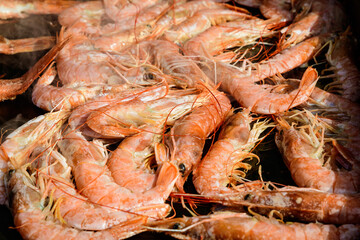 Close up of large portion of cooked shrimps on black grill at a street food festival, ready to eat healthy seafood, beautiful orange monochrome outdoor background.