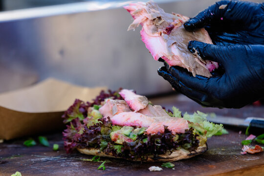 Hands of a man preparing a fresh healthy sandwich or burger with salmon guacamole and green salad on a brown wooden table at a street food market, side view photograph with soft focus .