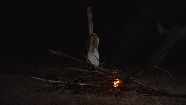 Hand of inquisitor in black shroud igniting bonfire with flaming torch with innocent hopeless woman in white dress tied to wooden pole, convicted as witch, outdoors at night during medieval witchhunt.