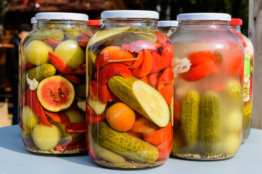 Three jars with mixed pickles on a white table for sale at a food market with pickled cucumbers, green tomatoes, red peppers, melon and cauliflower, side view with soft focus of tasty healthy food.