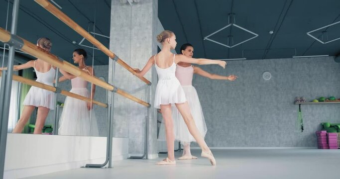 Female teacher shows ballet moves to preteen girl ballerina raising hand by wooden barre in studio at dance lesson slow motion