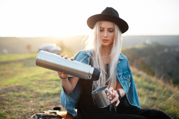 Portrait of young teenage blonde girl, pouring tea into the steel cup from thermos bottle. Outdoor background of sunset. Wearing black hat and denim jacket. - obrazy, fototapety, plakaty