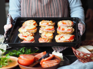 Elderly woman made hot sandwiches with cheese, tomatoes, sausage on toast and keeps them on a baking sheet from home oven.Healthy natural food. Home cooking