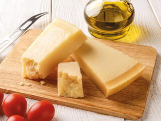 Wedge of italian hard cheese Parmesan or Grana Padano on a brown wood cutting boad near small red tomatoes, olive oil and cheese knife over white wood table. High key image.