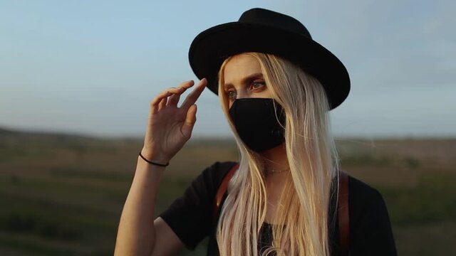 Outdoor portrait of young blonde girl wearing medical face mask against coronavirus and covid-19. Dressed in  black with hat.