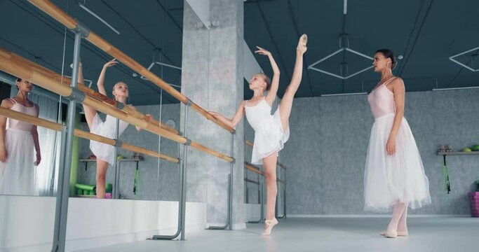 Concentrated girl ballerina does exercise with raised in splits leg leaning on barre under teacher control in studio slow motion