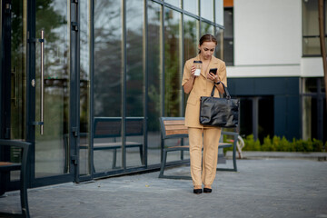 Smiling fashionable woman talking on mobile phone and drinking takeaway coffee in the city. Portrait Of Successful Business Woman Holding Cup Of Hot Drink In Hand On Her Way To Work On City Street