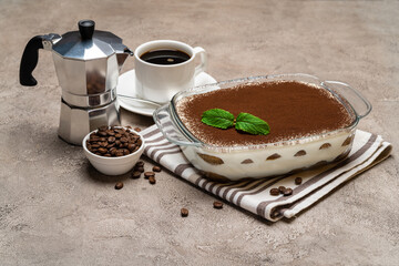 Traditional Italian Tiramisu dessert in glass baking dish, cup of espresso and mocha coffee maker on concrete background