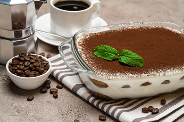 Traditional Italian Tiramisu dessert in glass baking dish, cup of espresso and mocha coffee maker on concrete background