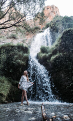 Vertical shot of an attractive young woman in a light summer dress posing with a waterfall behind