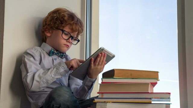 The Future Scientist. Auburn curly boy in glasses is sitting by the window and looking at the tablet. Near a large stack of books