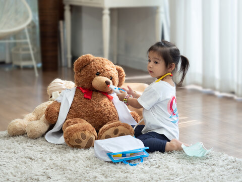 Cute little Asian girl playing doctor and using toy stethoscope with big brown teddy bear.