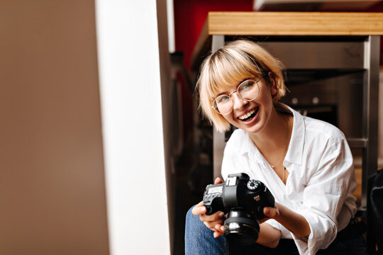 Laughing girl with short haircut holding camera. Indoor shot of happy female photographer in white shirt.