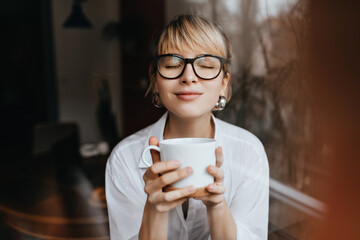 Front view of pretty woman enjoying coffee flavour with closed eyes. Smiling blonde girl in glasses holding cup of tea.