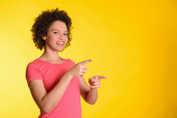 Young Brazilian woman smiling, on a yellow orange background, showing with both hands to the right, with space for text.