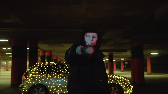 Man in scary white mask mask are holding a baseball bat and katana. Murderers at night in the parking lot against the backdrop of a car with a garland.