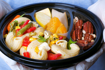 Safe food delivery. Pasta salad, slices of fruit and mix of nuts in the disposable bowl held by hands in gloves	