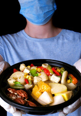 Safe food delivery. Pasta salad, slices of fruit and mix of nuts in the disposable bowl held by hands in gloves	