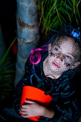 Portrait of Little girl in witch costume sitting in the night in the autumn park. Child with candy bucket in fall forest.