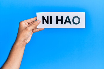 Hand of hispanic man holding chinese greeting ni hao word paper over isolated blue background.