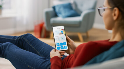 Hungry Young Woman is Lying on a Couch at Home and Using Smartphone for Ordering a Food via Advanced Food Delivery App with Modern User Interface. Perfect Life in Cozy Apartment