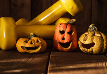Small ceramic Halloween pumpkins with two yellow dumbbells. Healthy fitness lifestyle autumn composition on wooden background.
