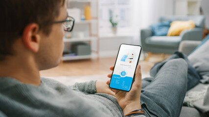 Over the Shoulder Shot of a Hungry Young Man Sitting on a Couch at Home and Using Smartphone for Ordering a Food via Advanced Modern Food Delivery App. Modern, Cozy and Bright Living Room.