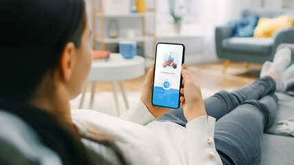 Over the Shoulder Shot of a Hungry Young Woman Sitting on a Couch at Home and Using Smartphone for Ordering a Food via Advanced Modern Food Delivery App. She is Waiting for Delivery.