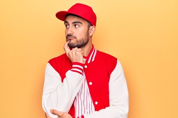 Handsome sporty man with beard wearing baseball jacket and cap over yellow background thinking...