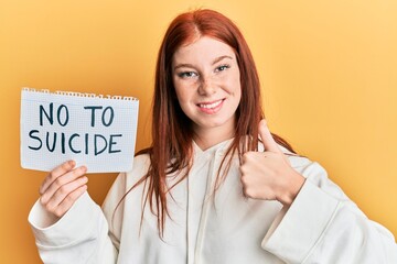 Young red head girl holding no to suicide paper smiling happy and positive, thumb up doing...