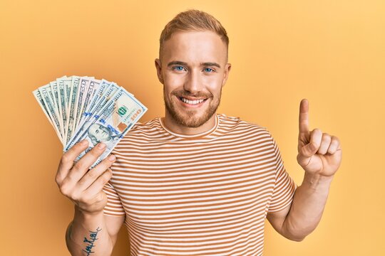 Young caucasian man holding dollars smiling with an idea or question pointing finger with happy face, number one