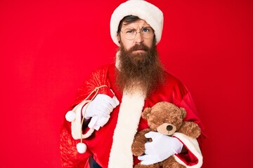 Handsome young red head man with long beard wearing santa claus costume holding teddy bear skeptic and nervous, frowning upset because of problem. negative person.