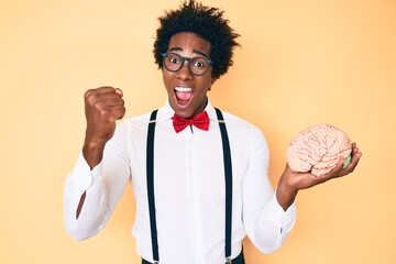 Handsome african american nerd man with afro hair holding brain screaming proud, celebrating...