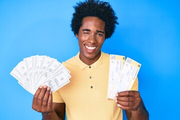 Handsome african american man with afro hair holding boarding pass and bunch of dollars winking...