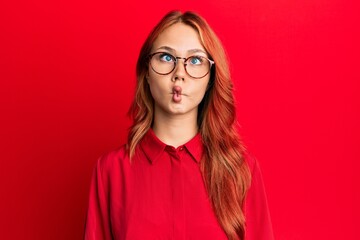 Young beautiful redhead woman wearing casual clothes and glasses over red background making fish face with lips, crazy and comical gesture. funny expression.