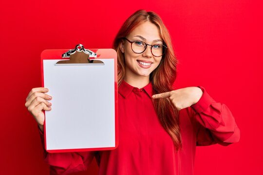 Young redhead woman holding clipboard with blank space pointing finger to one self smiling happy and proud