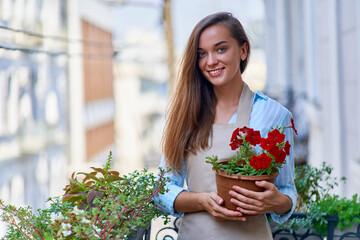 Portrait of happy smiling attractive woman gardener wearing apron holding flower pot on a balcony