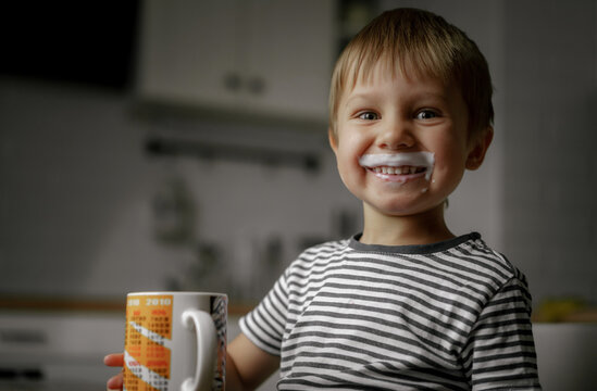 cute little boy with kefir mustache sitting at the table with cup in his hands and smiling. Image with selective focus