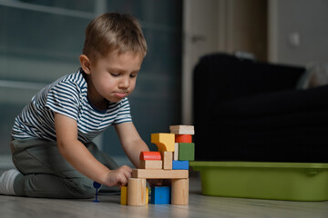 Naklejka premium Adorable little caucasian boy making funny face and playing with a tower out of colorful wooden blocks