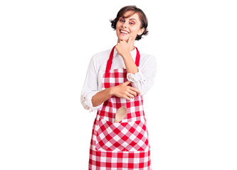 Beautiful young woman with short hair wearing professional cook apron looking confident at the camera with smile with crossed arms and hand raised on chin. thinking positive.