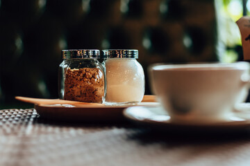Sugar in pot on wood plate near cup of coffee.