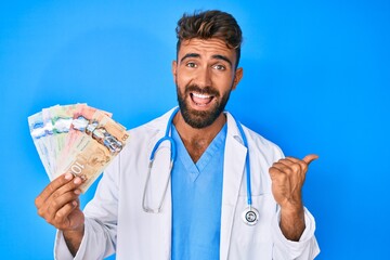 Young hispanic man wearing doctor uniform holding canadian dollars pointing thumb up to the side...