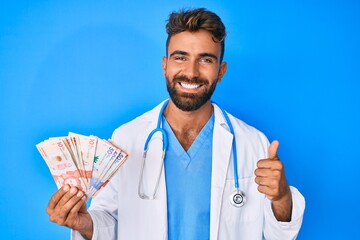 Young hispanic man wearing doctor uniform holding colombian pesos smiling happy and positive, thumb up doing excellent and approval sign