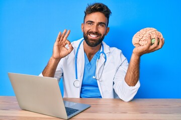 Young hispanic man wearing doctor uniform working at the clinic holding brain doing ok sign with...