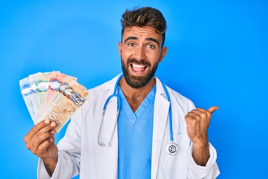 Young hispanic man wearing doctor uniform holding canadian dollars pointing thumb up to the side smiling happy with open mouth
