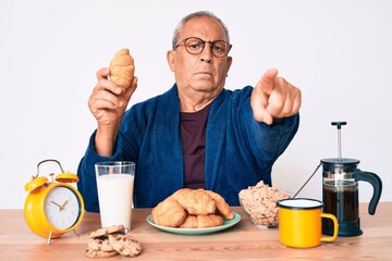 Senior handsome man with gray hair sitting on the table eating croissant for breakfast pointing with finger to the camera and to you, confident gesture looking serious