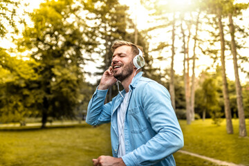 Smiling handsome young man singing with his arms raised as he uses headphones to listen to music in the park. Happy beautiful man enjoying life outdoors. Happiness, lifestyle concept. 