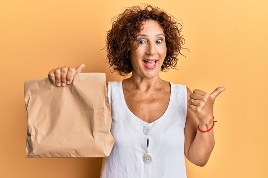 Beautiful middle age mature woman holding take away paper bag pointing thumb up to the side smiling happy with open mouth