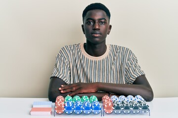 Young african american man sitting on the table with poker chips and cards with serious expression on face. simple and natural looking at the camera.