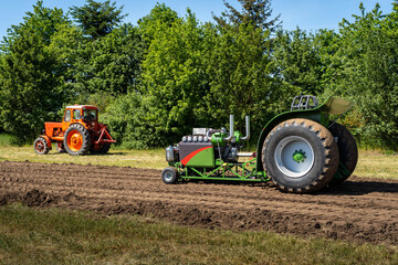 Green racing tractor with heavy back wheels in a field with trees in background, orange tractor in background